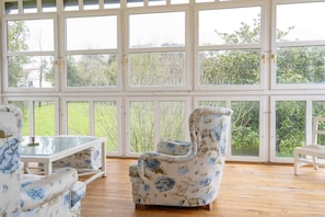 A conservatory with large glass doors, a floral armchair, a white chair, and a glass-top table.