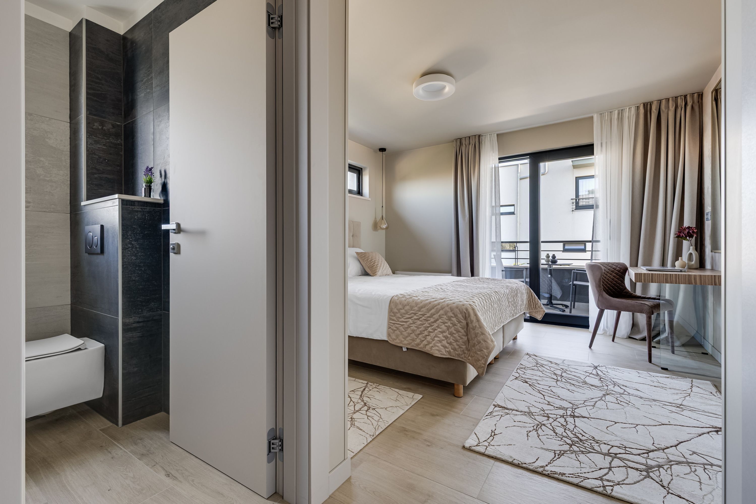 A modern hotel room with a bed, a desk, a chair, a bathroom, and a balcony with a view.