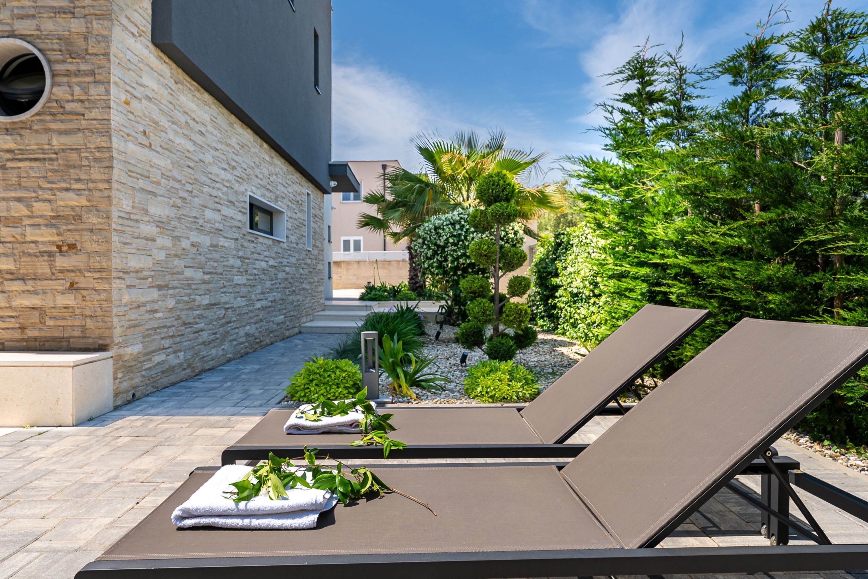 A modern outdoor lounge area with two reclining chairs, a stone wall, and a variety of greenery.