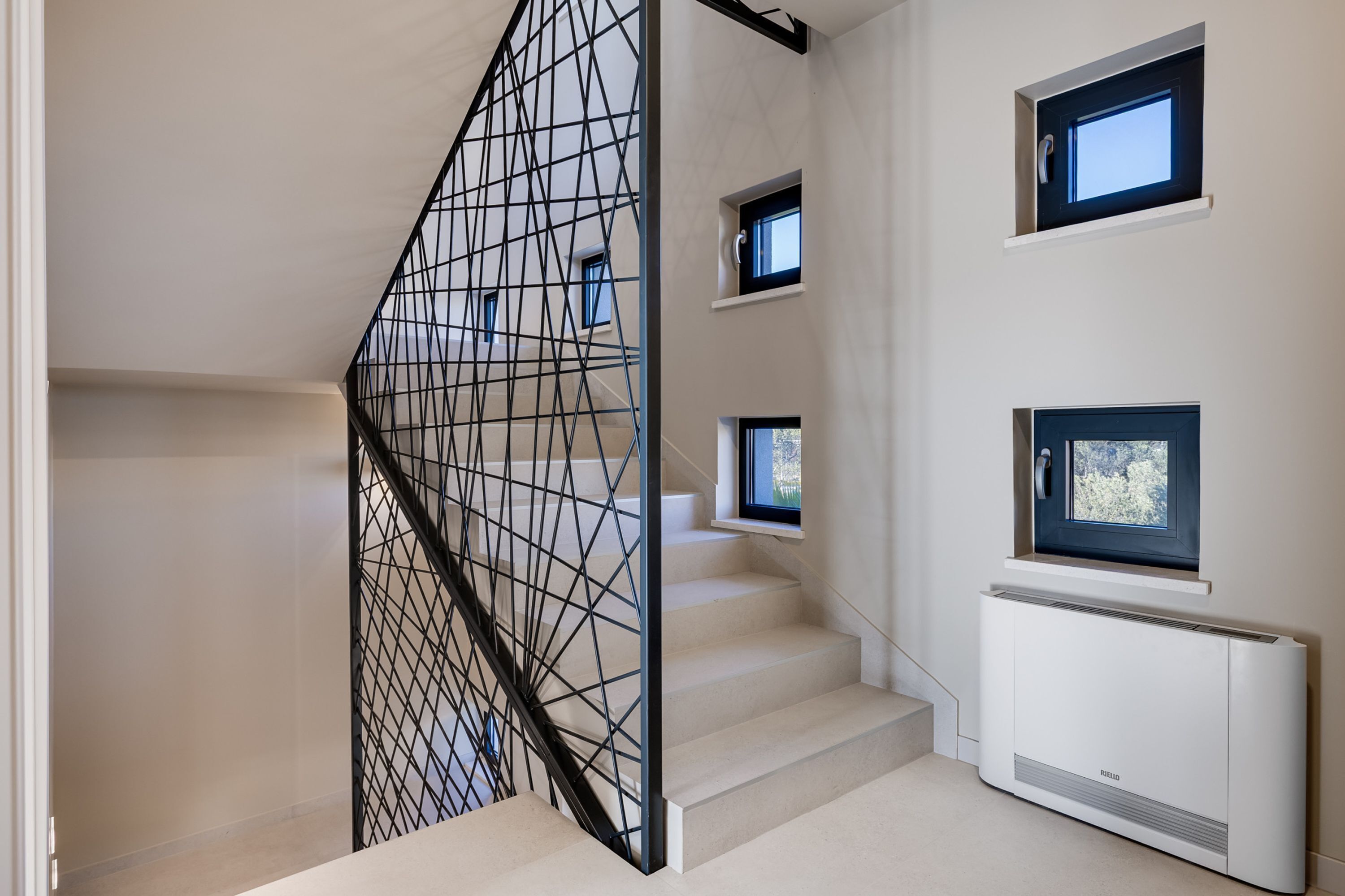 A staircase with a geometric metal railing, a white wall with multiple windows, and a white radiator.