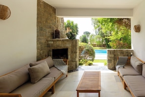 Ein gemütliches Wohnzimmer mit Kamin aus Stein, zwei Sofas, einem Holztisch und Blick auf einen Pool.