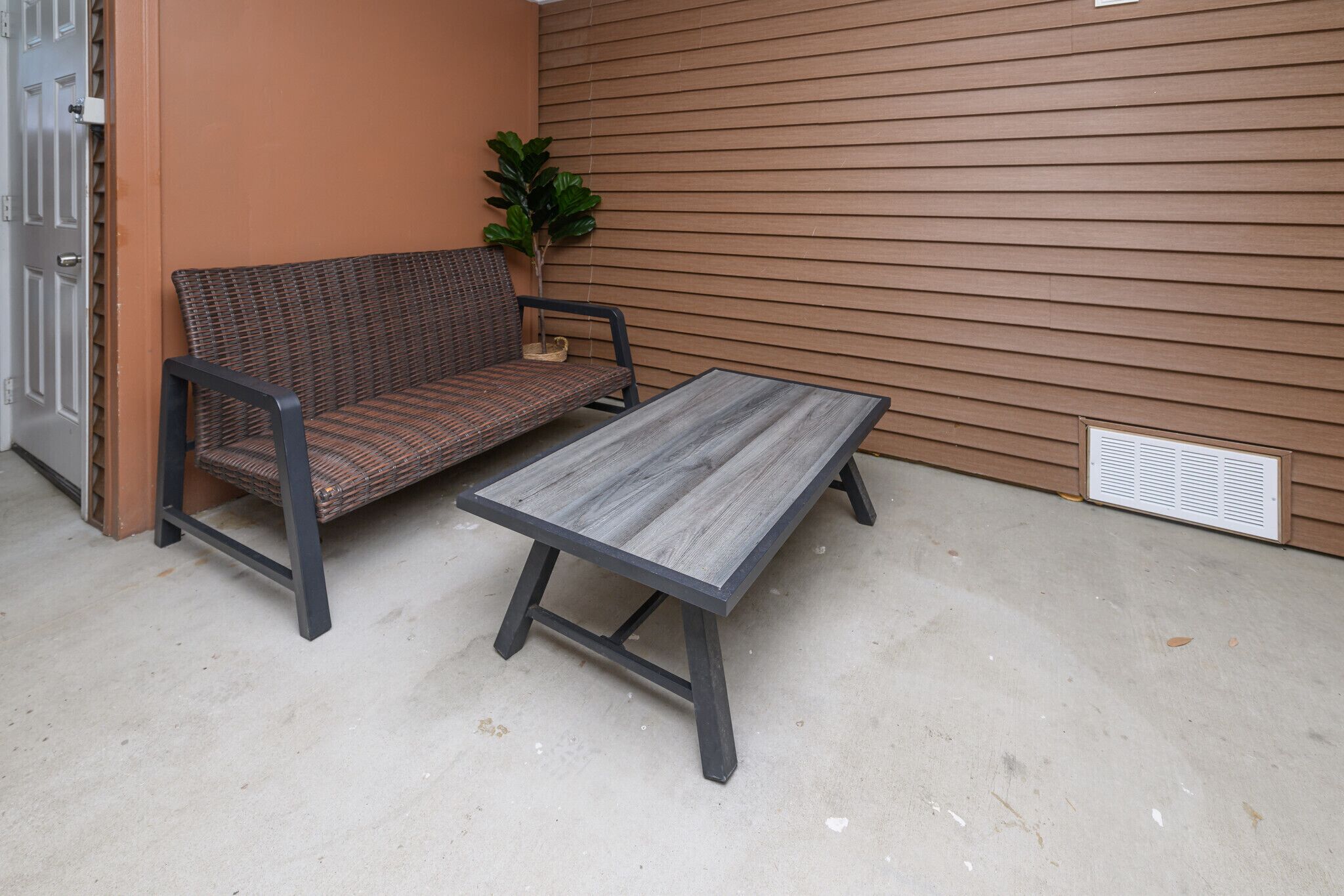 A concrete floor, a wicker sofa, a metal table, a potted plant, and a brown wall with horizontal siding.