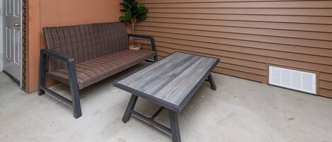 A concrete floor, a wicker sofa, a metal table, a potted plant, and a brown wall with horizontal siding.