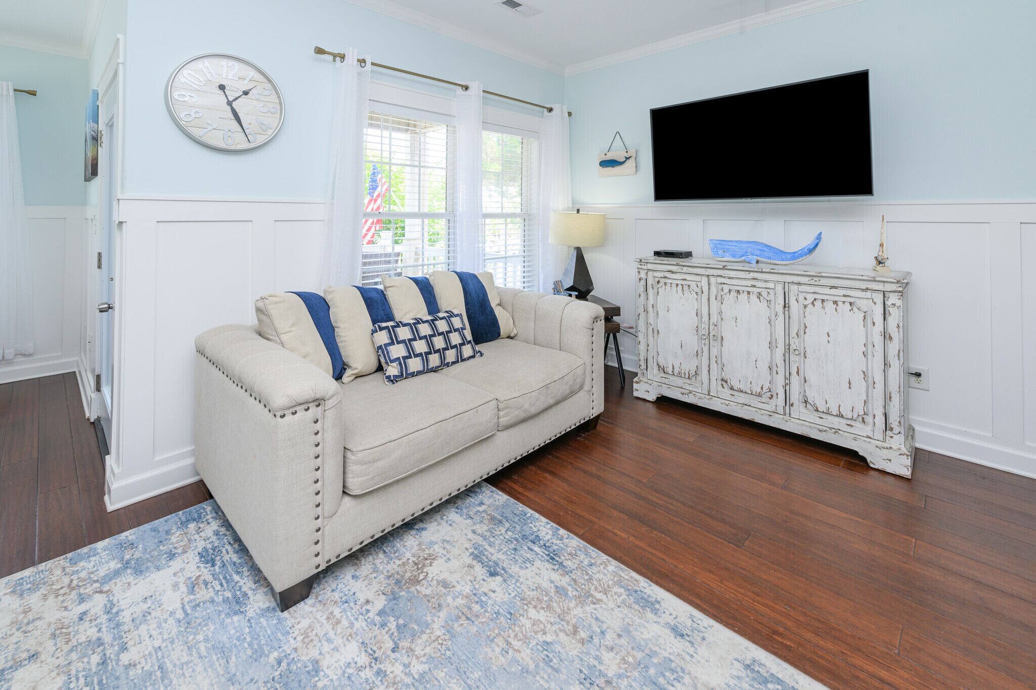 A living room with a beige sofa, a blue and white patterned throw, a wooden cabinet, a wall clock, and a flat-screen TV.