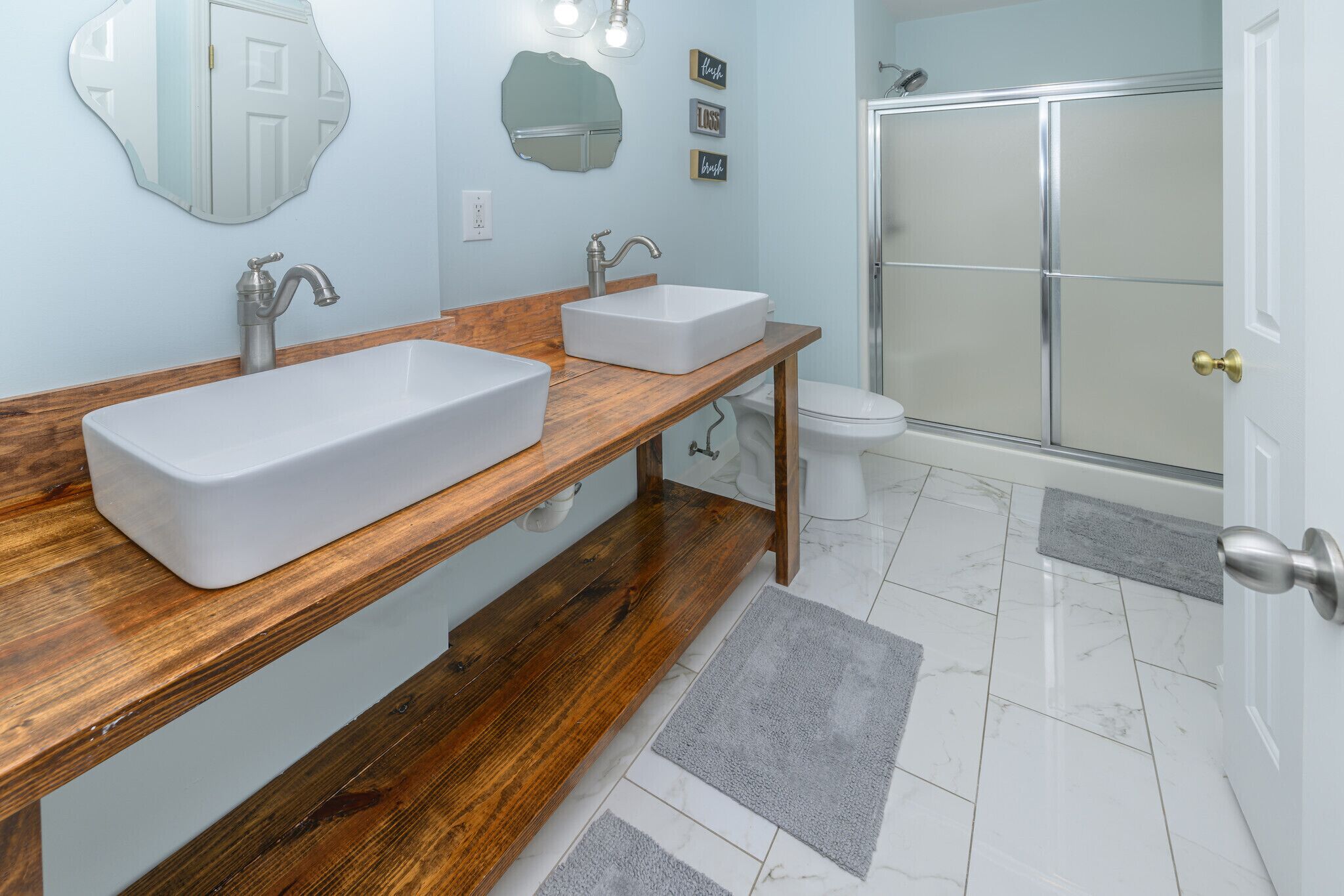 Un baño con dos lavamanos, un tocador de madera, un inodoro, una ducha y una puerta con pomo.