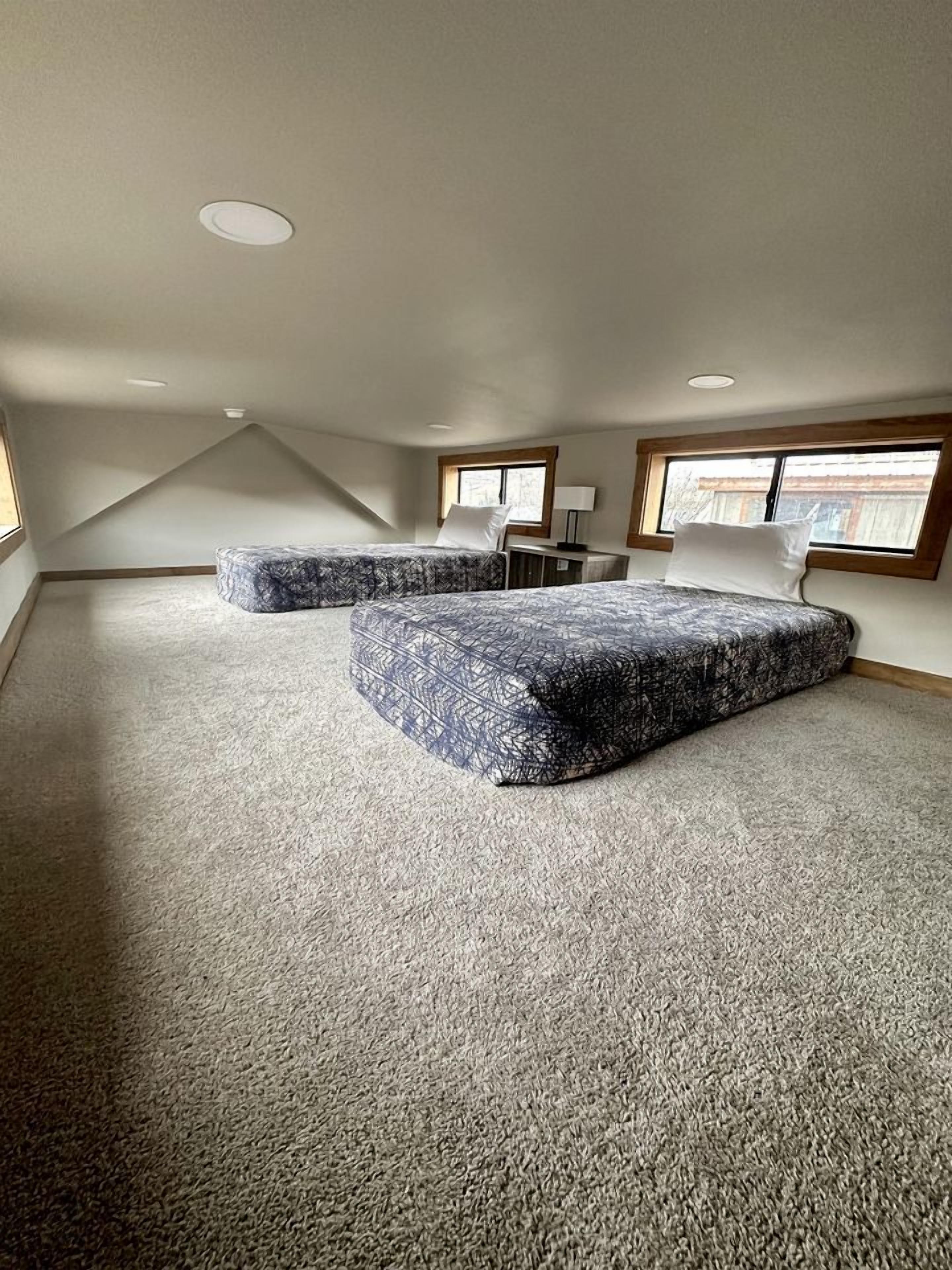 A room with a sloped ceiling, two large windows, a bed with patterned bedding, and a carpeted floor.