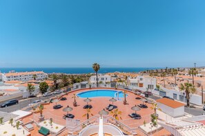 Ein Pool auf dem Dach mit Liegestühlen und Sonnenschirmen, umgeben von Gebäuden und Palmen, mit Blick auf die Küste.