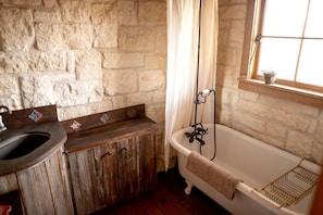 A rustic bathroom with a stone wall, wooden vanity, a bathtub, and a window with a plant.