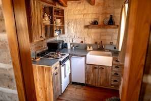 A compact kitchen with wooden cabinets, a stove, a sink, and a stone wall.