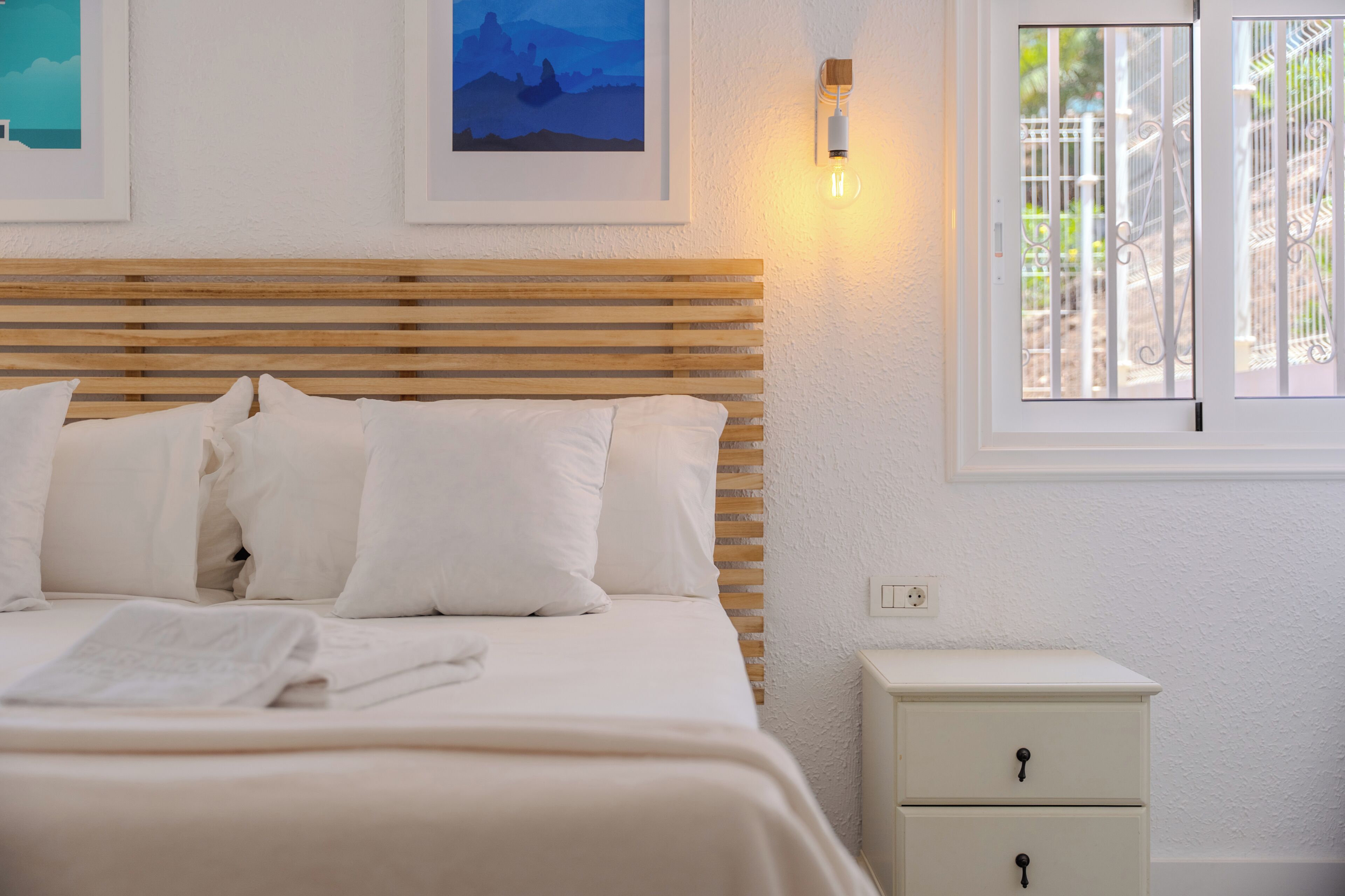 A bed with white bedding, a wooden headboard, a bedside table with drawers, and a window with a view of a white fence.