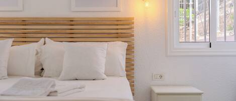 A bed with white bedding, a wooden headboard, a bedside table with drawers, and a window with a view of a white fence.