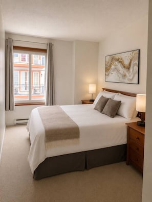 Ein Hotelzimmer mit einem großen Bett, zwei Nachttischen, einem fenster mit Vorhängen, einem an der Wand hängenden abstrakten Gemälde und Blick auf Gebäude.