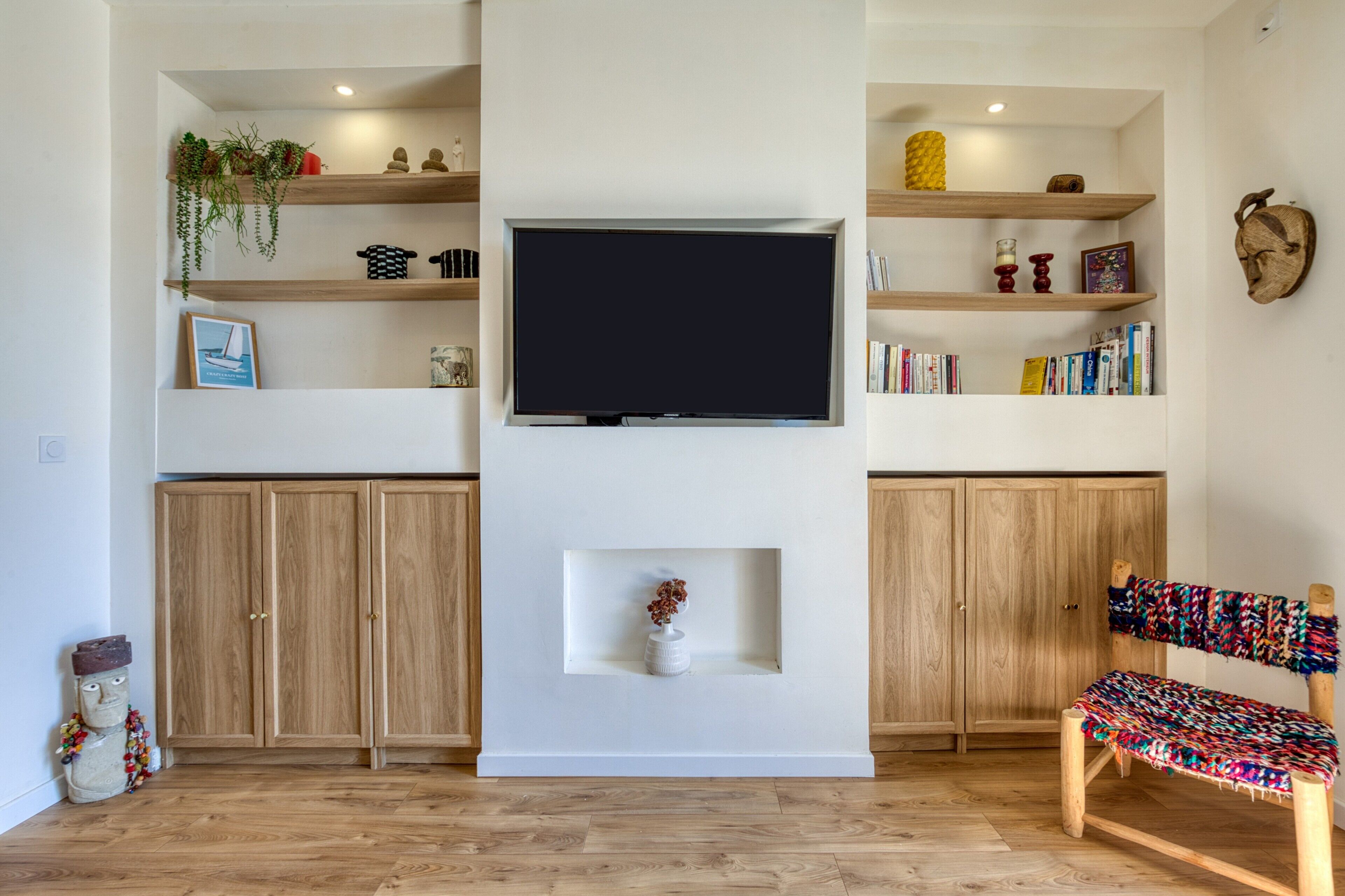 A living room with a flat-screen TV, wooden shelves, a bookshelf, a cabinet, a decorative wall piece, and a woven chair.