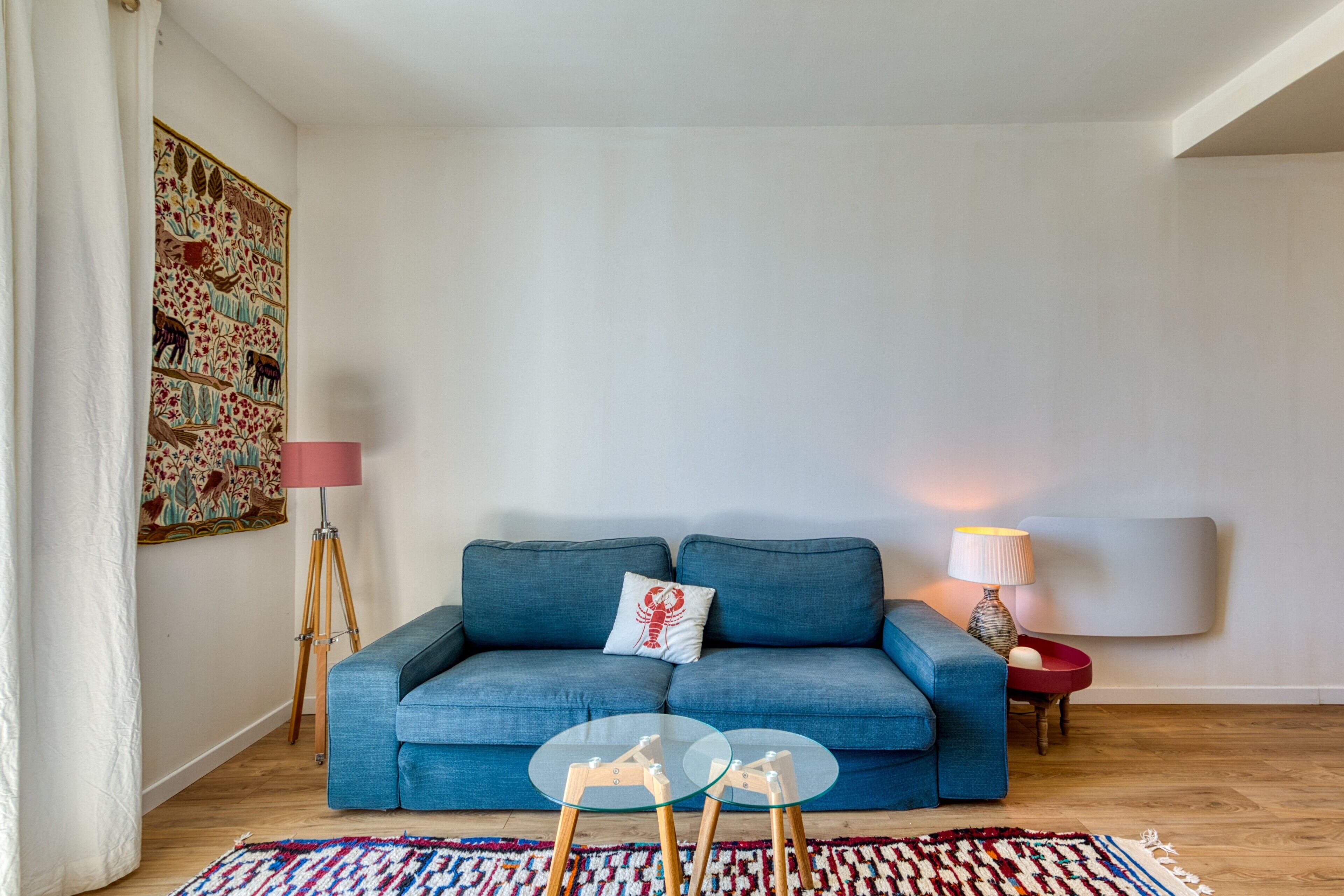 A living room with a blue sofa, a glass coffee table, a patterned rug, a floor lamp, and a wall-mounted lamp.