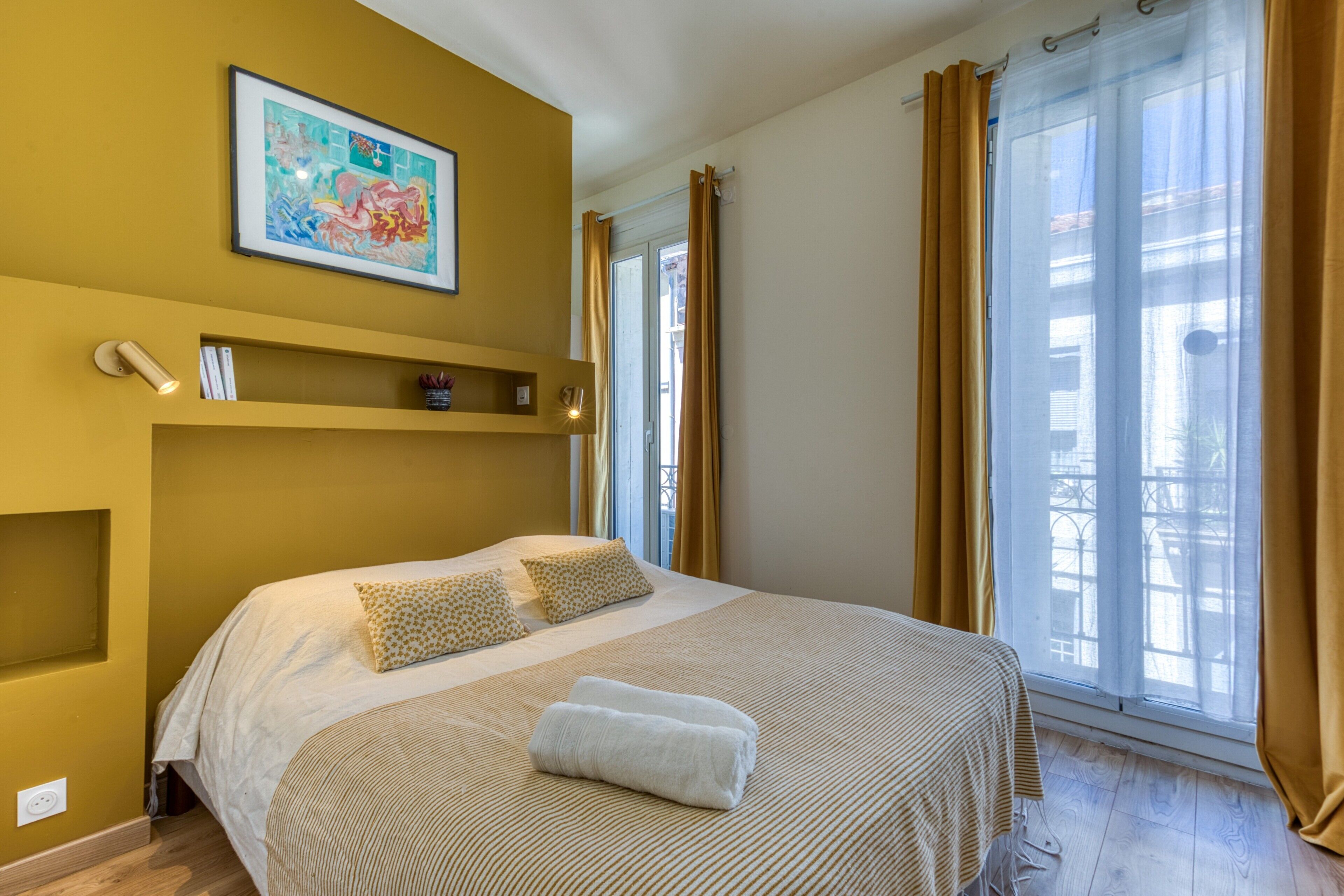 A hotel room with a bed, a painting on the wall, a shelf with a phone and books, and a window with curtains.