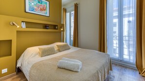 Quarto de hotel com uma cama, um quadro na parede, uma estante com um telefone e livros, e uma janela com cortinas.