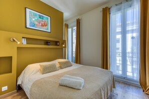 Hotelkamer met een bed, een schilderij aan de muur, een plank met telefoon en boeken, en een raam met gordijnen.