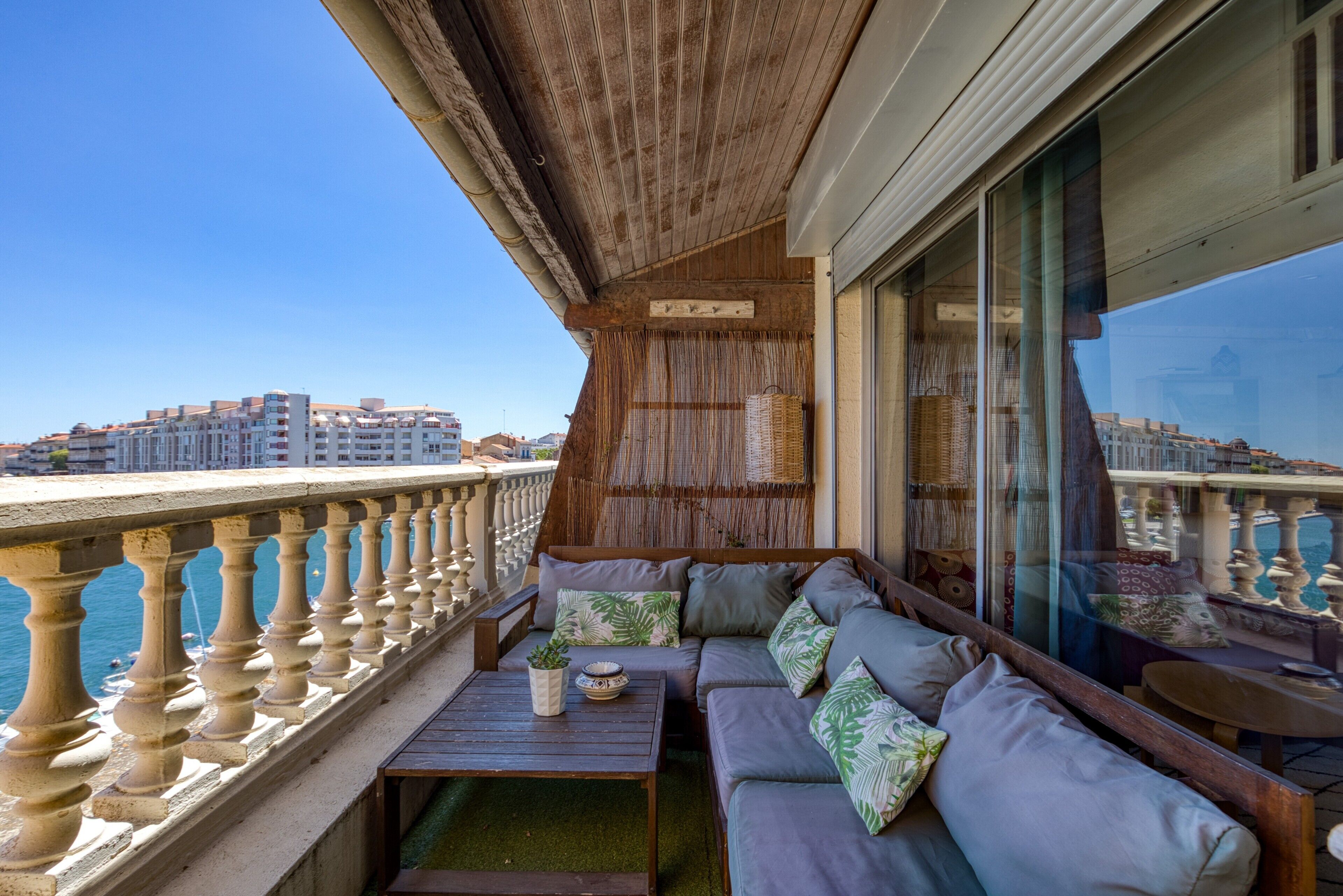 Un balcone con ringhiera in legno, un divano con cuscini, un tavolino con una pianta e vista sulla città.