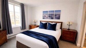 A bedroom with a large bed, two bedside tables, a dresser, a window with curtains, and a wall-mounted picture. - Heart of Tremblant - Johanssen, Place St. Bernard (Mont-Tremblant)