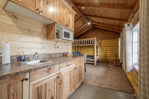 L’intérieur d’une cabane en bois comprend un coin cuisine, des lits superposés et une fenêtre avec des rideaux.