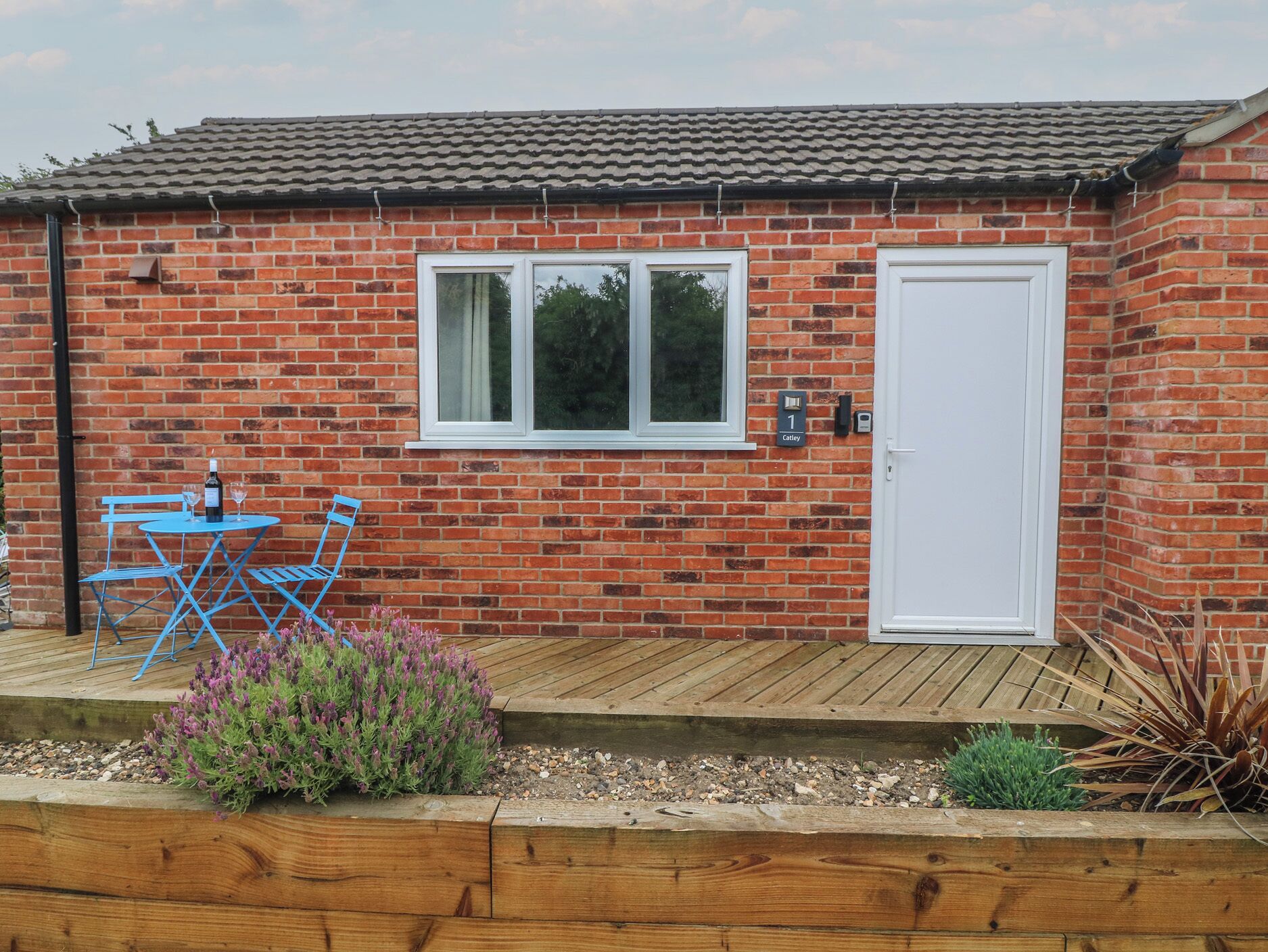 A brick house with a white door and windows, a small patio with blue chairs and a table, and a wooden deck.