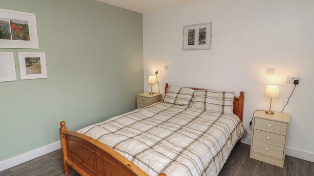 A bedroom with a wooden bed, two bedside tables with lamps, a small dresser, and framed pictures on the wall. - Walcott Lodges - Catley (Lincoln)