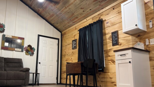 A room with a wooden ceiling, a white door, a brown sofa, a small table with chairs, and a white cabinet.