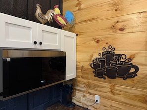 Una cocina con un microondas negro grande, armarios blancos y una pared de madera con un mural alusivo al café.