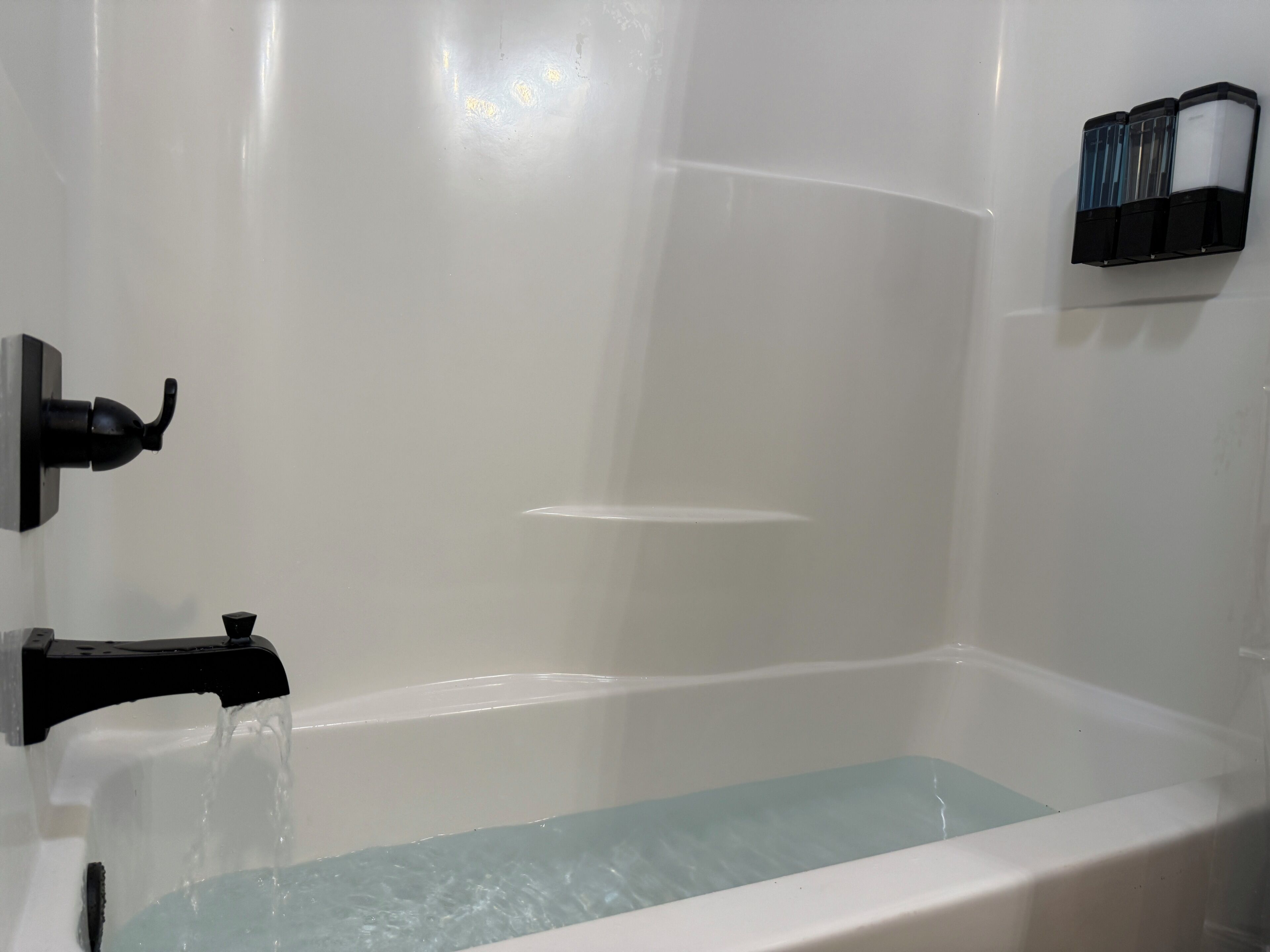 A bathtub with running water, a black faucet, and a wall-mounted soap dispenser.