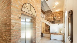 Un pasillo amplio con pared de ladrillo, un ascensor moderno, una escalera con barandillas de hierro forjado y una consola de madera.