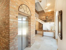 A spacious hallway with a brick wall, a modern elevator, a staircase with wrought iron railings, and a wooden console table.