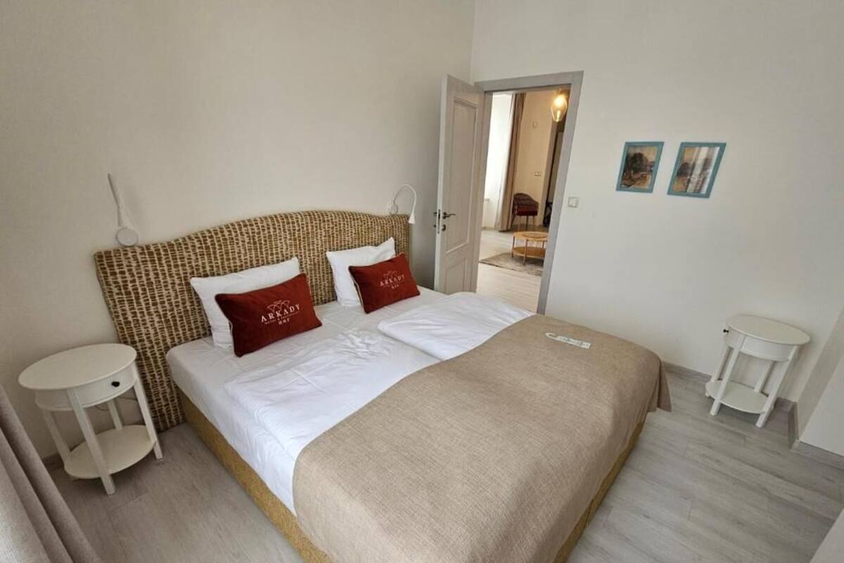 Un dormitorio ordenado con una cama, dos mesitas de noche, una puerta que da a otra habitación y una pequeña mesa blanca.