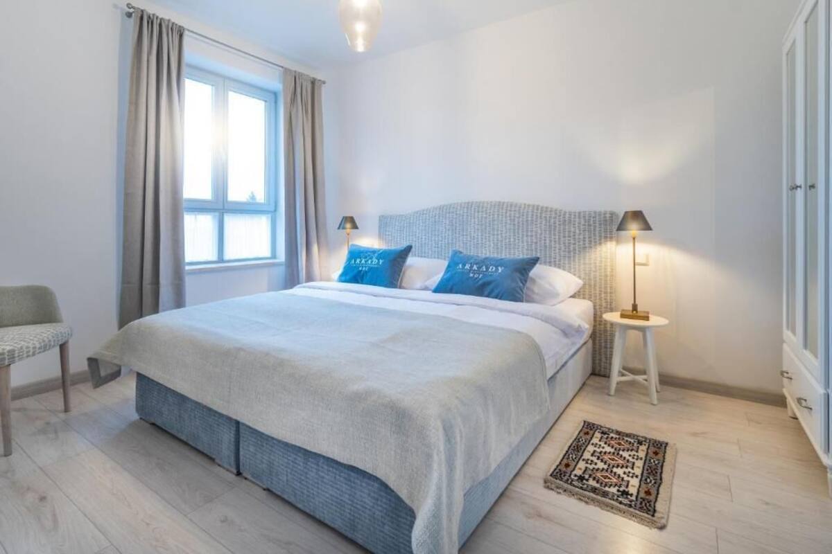 Un dormitorio con una cama, dos lámparas de noche, una silla, una ventana con cortinas y una alfombra estampada.