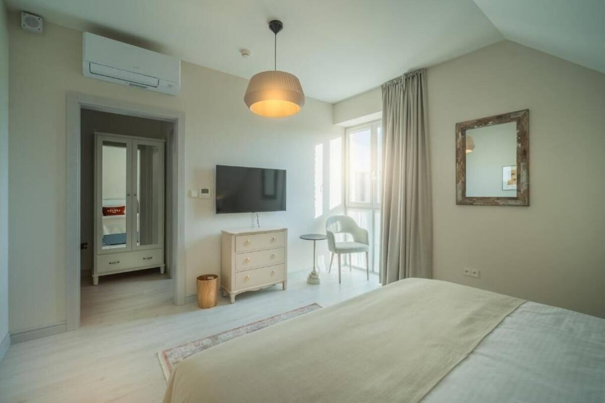 Un dormitorio con cama, televisor, cómoda, silla, espejo y ventana con cortinas.