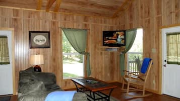 Quarto com paredes de madeira, um sofá, uma mesa, uma cadeira de balanço e uma televisão.