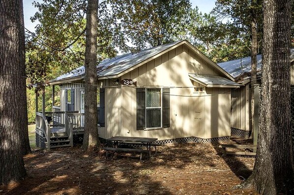 A small, single-story cabin with a porch, surrounded by trees.