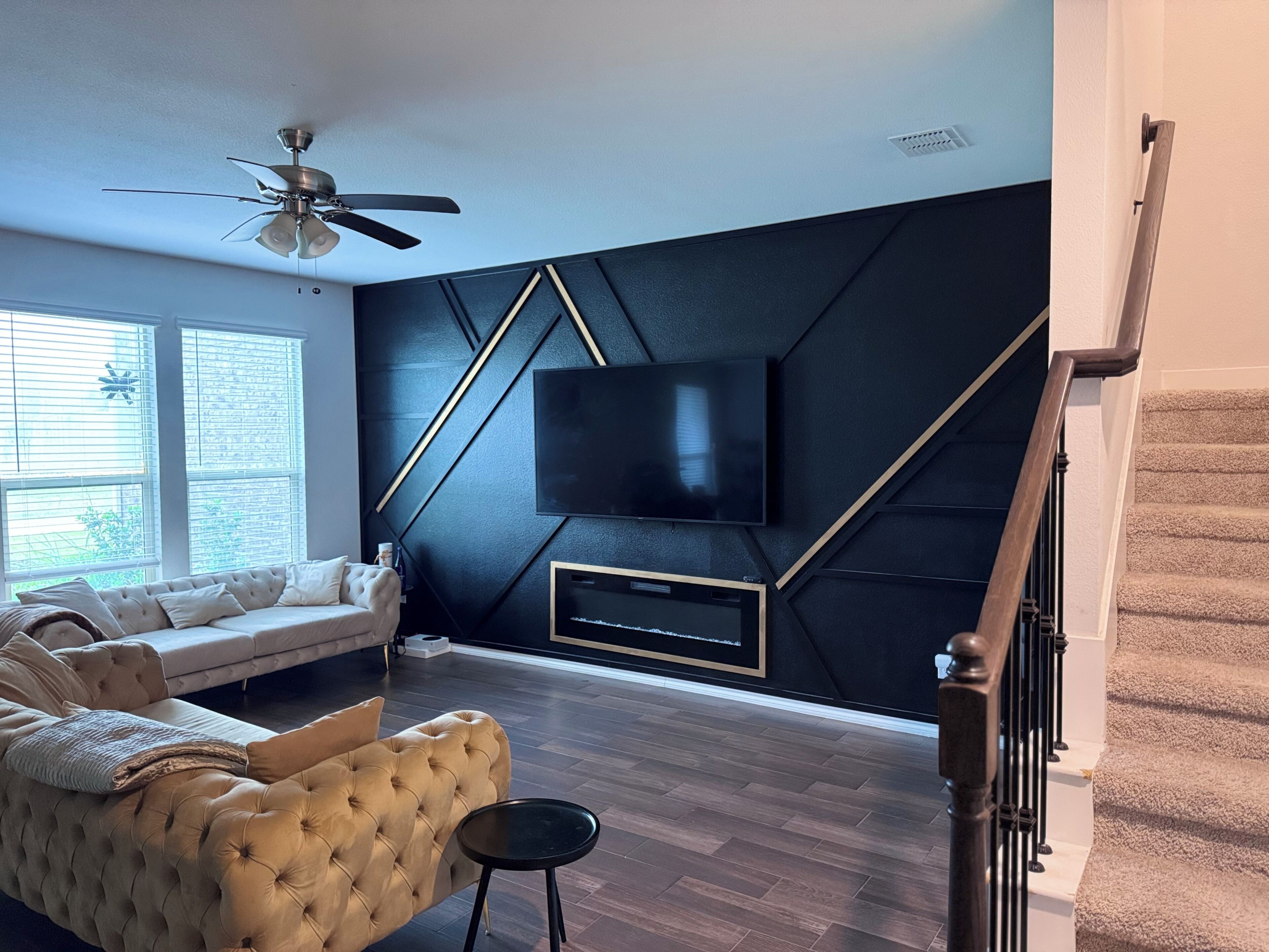 Un salon moderne avec un canapé, un téléviseur fixé sur un mur noir, un ventilateur de plafond et un escalier.
