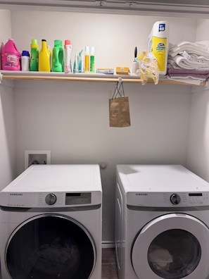 Un espace buanderie comprenant deux machines à laver Samsung, une étagère avec des produits de nettoyage et un sac de courses suspendu.