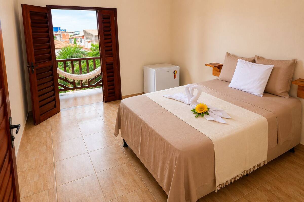 Habitación de hotel con cama, un pequeño electrodoméstico blanco, una puerta de madera y vistas de un jardín a través de una puerta abierta.