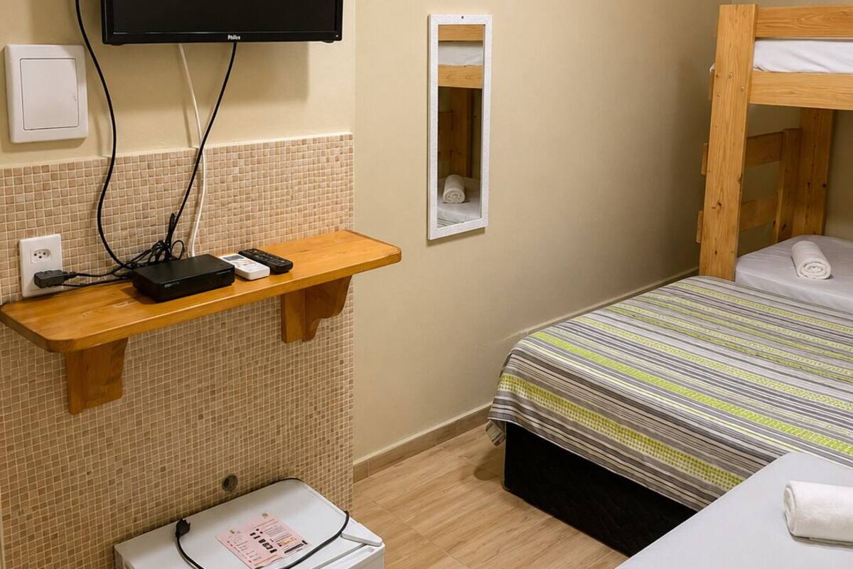 Habitación de hotel con una cama, un televisor instalado en la pared, un refrigerador pequeño y un estante de madera con un distribuidor de energía.