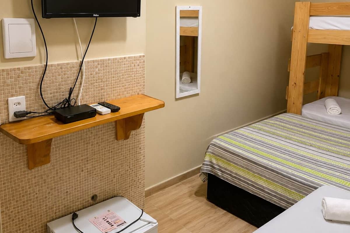 A hotel room with a bed, a TV mounted on the wall, a small refrigerator, and a wooden shelf with a power strip.