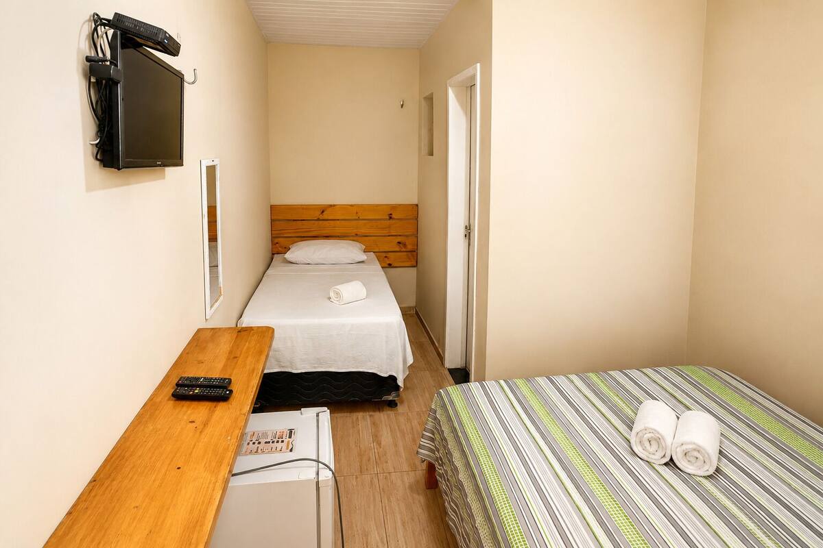 Una habitación de hotel pequeña con una cama individual, un escritorio con computadora y teclado, un televisor montado en la pared y un refrigerador pequeño.