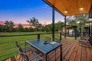 A wooden deck with a table and chairs, a covered area with a view of the sunset, and a building with glass windows. - Family-Friendly Tiny Home Resort with Pool, Goats & Pond in Hempstead near Houston (Hempstead)