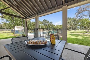 A covered patio with a table set for a meal, a grill, and a view of a park. - Family-Friendly Tiny Home Resort with Pool, Goats & Pond in Hempstead near Houston (Hempstead)