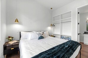 A modern bedroom with a large bed, bedside tables, a nightstand, a window with blinds, and a door leading to another room. - Family-Friendly Tiny Home Resort with Pool, Goats & Pond in Hempstead near Houston (Hempstead)