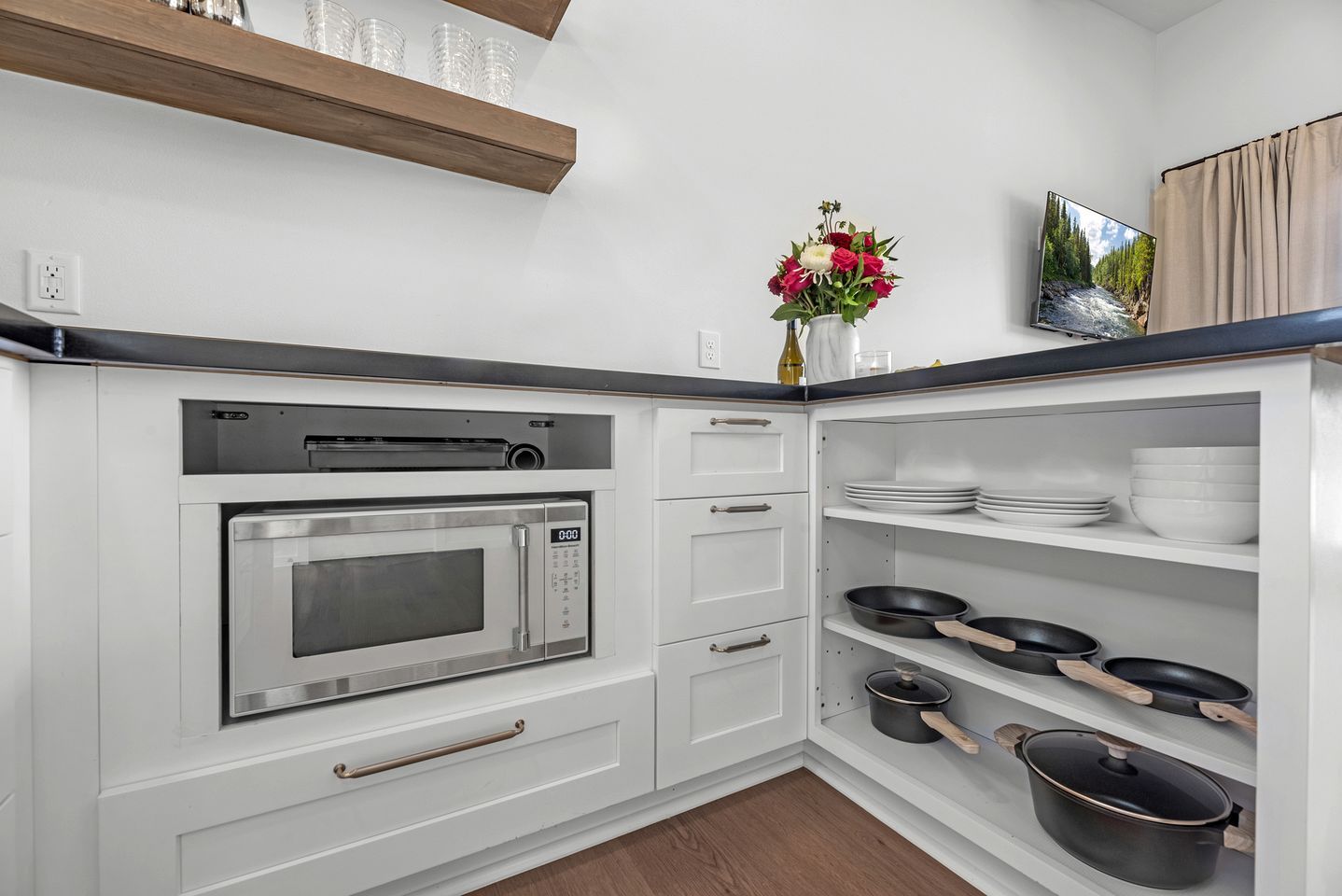 A kitchen with a built-in microwave, white cabinets, a shelf with dishes, and a vase with flowers.