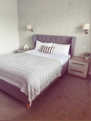 A bedroom with a bed, two bedside tables, a nightstand, a lamp, and a wall-mounted light fixture. - Malva - Modern 2 Bedroom Bungalow with Free Private Parking on the NC500 (Wick)