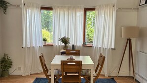 Een eetruimte met een tafel, stoelen, een lamp en uitzicht op groen door ramen.