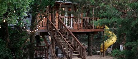 Une cabane dans les arbres avec un escalier en bois et un toboggan jaune, entourée d’un feuillage dense.