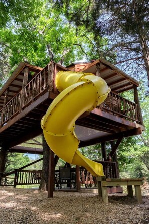 A wooden structure with a yellow slide, surrounded by trees. - Enchanting Forest Treehouse Hideaway with Farm Activities near Bingham Creek in Forestburg, Texas (Forestburg)